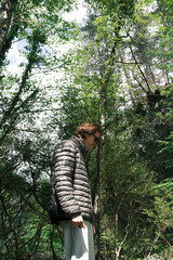 A young man wearing a gray puffer jacket stands in a sunlit forest on Mount Olympus, surrounded by dense greenery and trees.