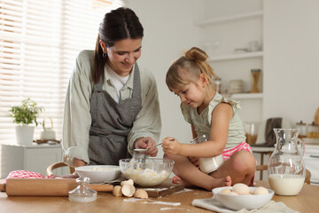 Little girl helping her mom making dough at table in kitchen