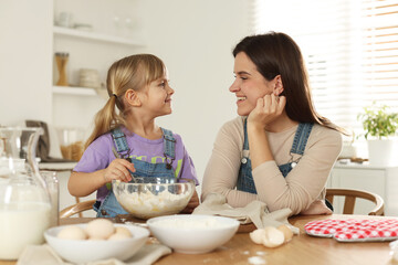 Little girl helping her mom making dough at table in kitchen