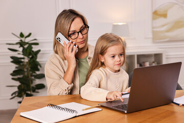 Work-family balance. Single mother talking by smartphone and her daughter drawing at wooden table...