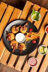 Stuffed prawns served with creamy white dip and fresh salad in a black plate placed on a rustic wooden crate, styled with lemon slices and red sauce in small white bowls on a warm brown background.