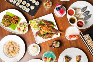 Top-down view of a diverse spread of dishes on a table, including fried mackerel, sushi, dim sum, kebab, chicken wings, pasta, two desserts, and two colorful cocktails in a restaurant setting