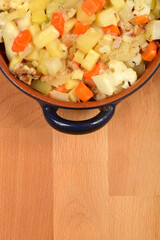A close-up, high-angle shot captures a rustic blue ceramic baking dish brimming with a savory mixture of chicken, potatoes, carrots, and cauliflower, resting on a wooden surface