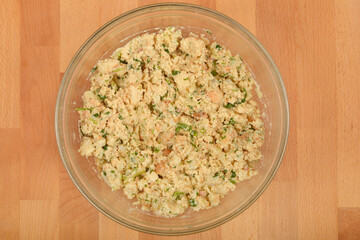 An overhead shot presents a glass bowl filled with a thoroughly mixed preparation for bread dumplings, featuring moist bread, chopped herbs, and other binding ingredients, all on a wooden surface