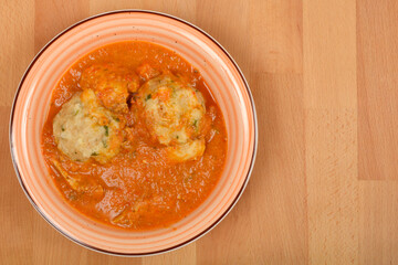 Overhead shot shows two bread dumplings submerged in a rich chicken paprikash with sauce, served in a bowl on a wooden surface