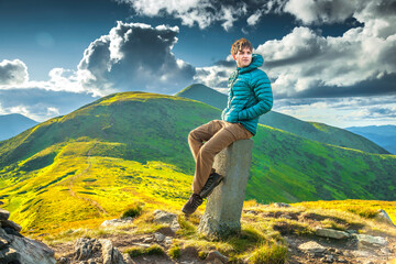 Man resting on a mountain peak in soft sunlight, wearing a green jacket and brown hiking pants,...