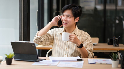 Happy businessman talking on phone and drinking coffee at desk.