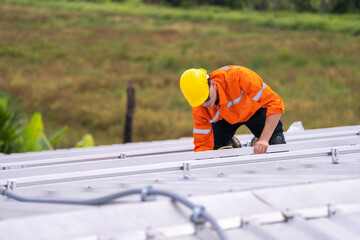 Worker in an orange safety uniform and helmet carefully inspecting a metal rooftop under natural daylight. Depicts diligence, precision, and professional commitment in construction and maintenance.