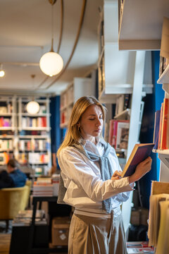 Interested thoughtful woman browsing educational books in bookshop, standing by window. Focused elegant middle-aged female carefully reads content of selected novel bestseller, shopping in bookstore. 