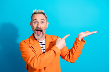 Naklejka premium Vibrant mature man in orange jacket presenting on a blue background with expressive gestures and joyful expression