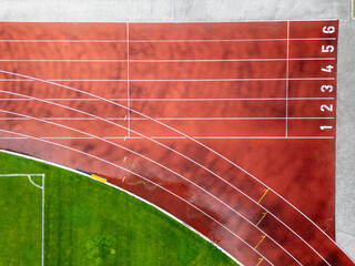Aerial view of a red running track with marked lanes and curved lines. Athletic stadium section with part of a green football field. Sports and fitness concept.