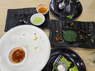 Rectangular and circular black and white food plates on dining table. Plate with some food scraps left after eating. Vegetable leaves, dipping sauce, chili left on the plate.Eat all food on your plate