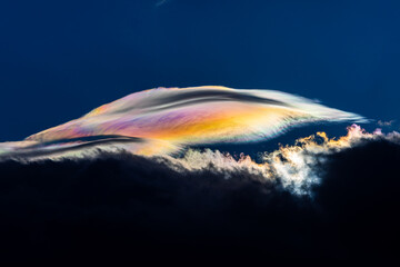 Spectacular iridescent cloud glows in vivid rainbow colors above dark storm clouds. Rare optical weather phenomenon captured against a deep blue sky. Amazing type of clouds.