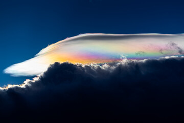 Spectacular iridescent cloud glows in vivid rainbow colors above dark storm clouds. Rare optical weather phenomenon captured against a deep blue sky. Amazing type of clouds.