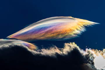 Spectacular iridescent cloud glows in vivid rainbow colors above dark storm clouds. Rare optical weather phenomenon captured against a deep blue sky. Amazing type of clouds.