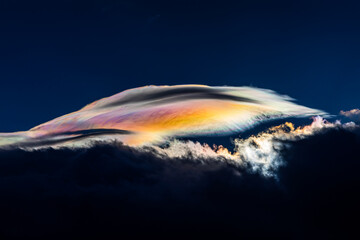 Spectacular iridescent cloud glows in vivid rainbow colors above dark storm clouds. Rare optical weather phenomenon captured against a deep blue sky. Amazing type of clouds.