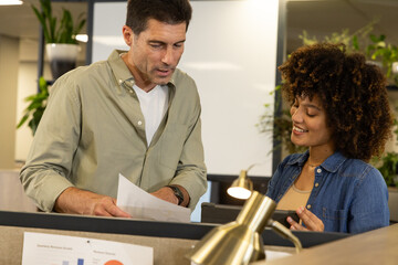 Diverse coworkers reviewing printed charts in office cubicle, with tablet and brass desk lamp