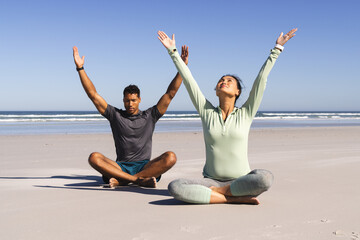 Fototapeta premium Couple practicing yoga on beach, meditating with arms raised under clear sky