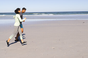 Fototapeta premium Diverse couple jogging along sandy beach at sunrise, embracing calm ocean breeze, copy space