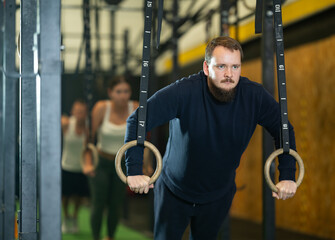Determined young guy working out with gymnastic rings in gym during weight training classes