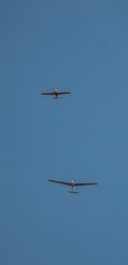 Two gliders are seen soaring through a clear, vast blue sky. One glider is positioned higher and...