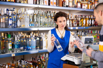 Adult saleswoman in uniform sells bottle of vodka to adult male customer in grocery store