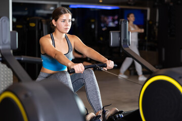 Fototapeta premium Determined young girl training on cable row machine in large fitness hall