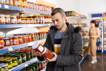 Man in store of imported Russian goods chooses canned lecho. Male buyer is looking for traditional national Russian grocery snacks lecho – stewed salad made from bell pepper, tomato sauce, onion