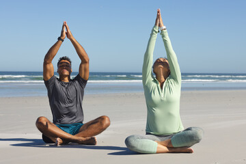 Fototapeta premium Diverse couple meditating on beach, raising hands over soft sand with gentle waves