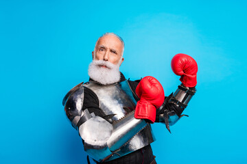 Senior man in medieval armor and boxing gloves posing against blue background