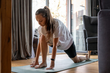 Woman performs a deep lunge stretch while practicing yoga on a mat in her living room. She focuses...