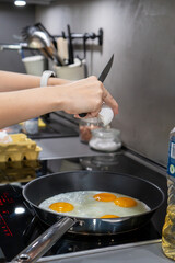 Hands are shown carefully cracking a fresh egg with a knife over a hot frying pan, adding it to other eggs already cooking sunny-side up for a delicious homemade meal.