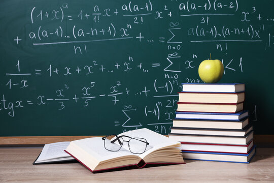 Books, glasses and apple on wooden table near blackboard with formulas