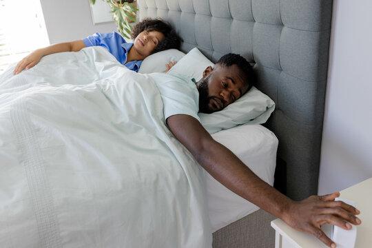 African American man reaching for alarm clock while waking up next to partner