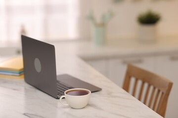 Laptop and cup of hot drink on white marble table in kitchen, selective focus