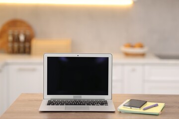 Fototapeta premium Laptop with blank screen, smartphone and stationery on wooden table in kitchen. Mockup for design