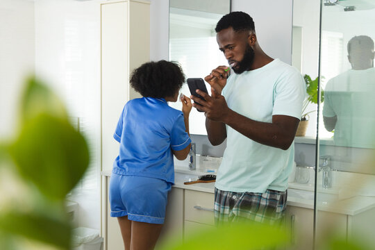 Young couple brushing teeth at sink in modern bathroom, holding smartphone and toiletries - Powered by Adobe