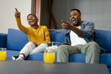 African American father and son laughing and pointing while watching TV on blue sofa