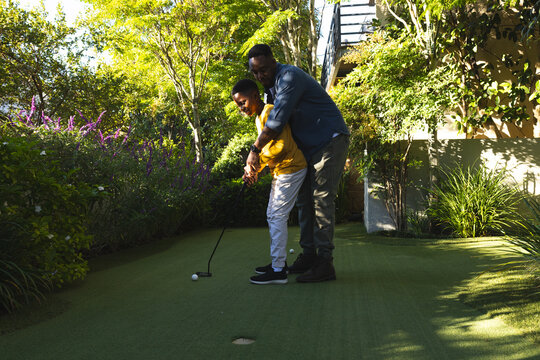 African American father and son coaching golf on green backyard lawn, holding golf club and ball - Powered by Adobe