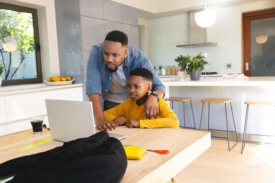 Father helping son with homework on laptop at kitchen table, both concentrating