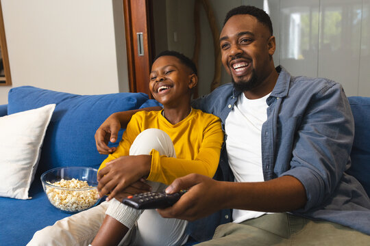 African American father and son watching TV on couch, laughing and enjoying popcorn