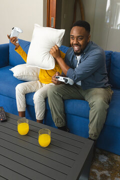 African American father and boy sitting on couch in living room, playing with controller and pillow