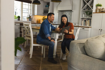 Mature Asian couple at home using tablet, smiling and enjoying time together in kitchen