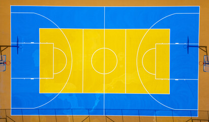 Aerial view of a colorful outdoor basketball court with vibrant blue and yellow markings, two hoops, and symmetrical layout. Shot from directly above.