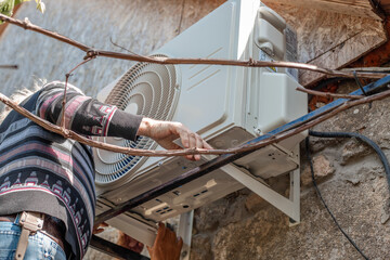 A man installs an outdoor air conditioner unit on iron mounting brackets on the wall of a stone...