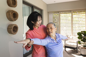 Asian woman helping elderly senior mother with stretching exercises at home, both smiling