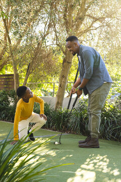 African American father and son playing mini golf outdoors among trees with putter and golf ball