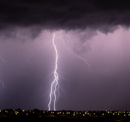 Single vertical lightning bolt pierces a purple stormy sky above a glowing urban skyline during a...