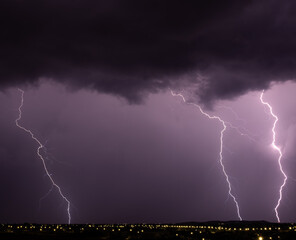 Multiple lightning strikes hit the earth under dramatic purple storm clouds above a city full of...