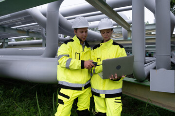Workers collaborate on a laptop beside industrial piping in a safety zone during daytime